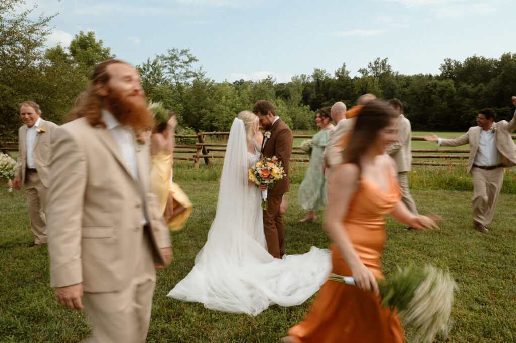 Bride and groom sharing a moment while wedding party moves around them in a grassy field