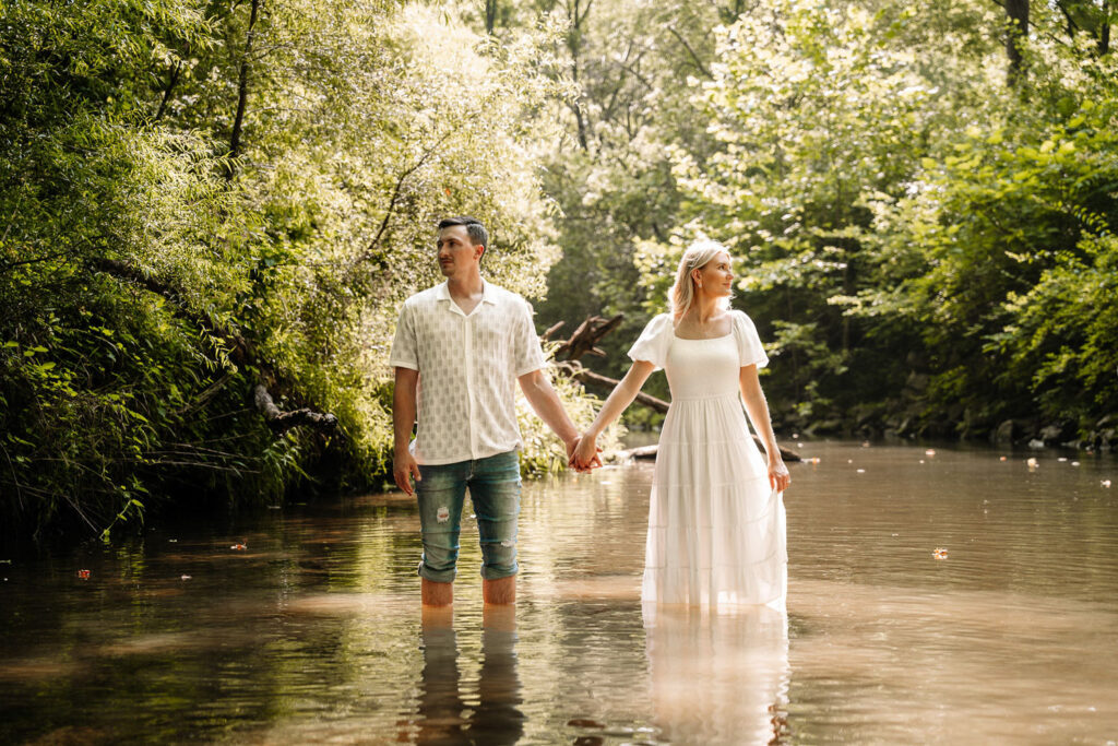 Couple standing in a shallow stream, holding hands amidst serene forest surroundings