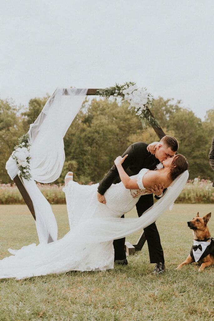 Bride and groom sharing a romantic dip under a floral wedding arch with their dog nearby