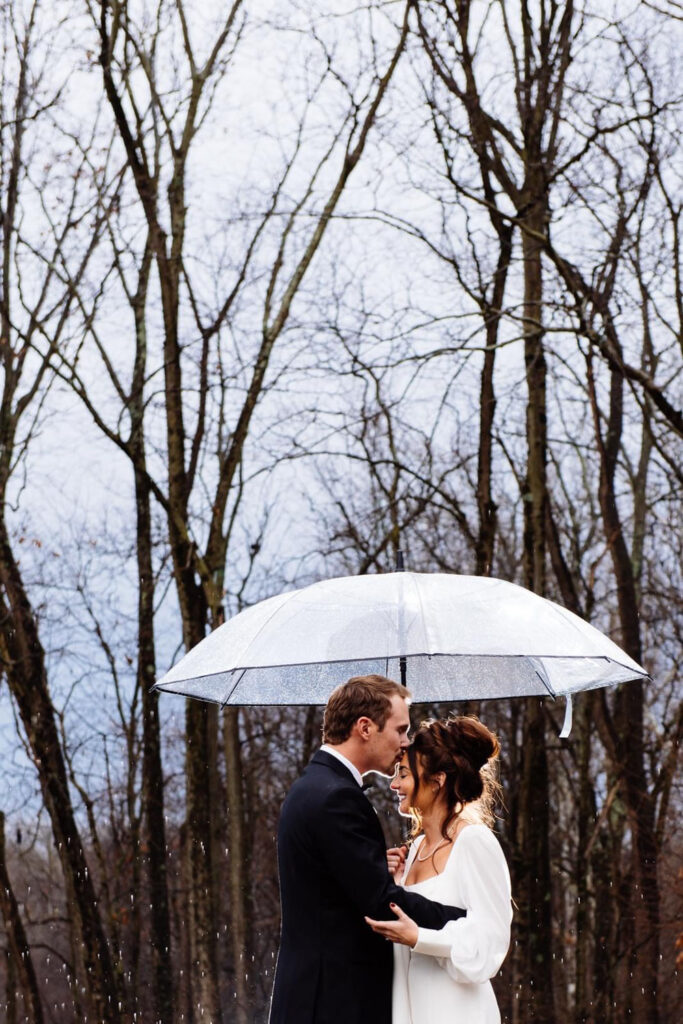 Couple sharing a kiss under a clear umbrella in a wooded area at a wedding reception venue in Indiana