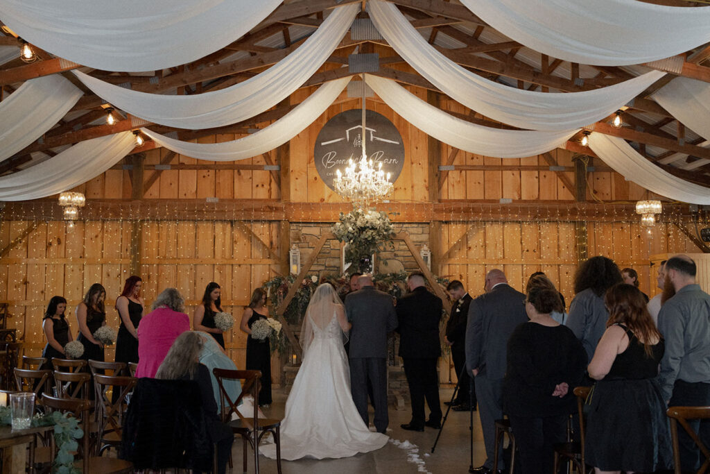 Indoor barn wedding ceremony with draped white fabric and string lights at a wedding reception venue in Indiana