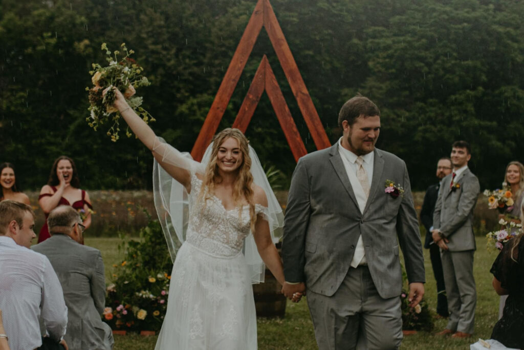 Bride raising her bouquet in celebration as she walks with her groom under a wooden wedding arch