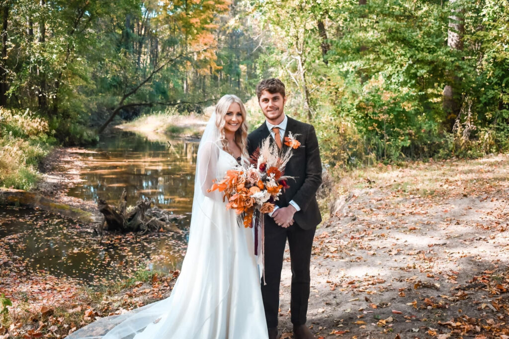 Bride and groom posing with vibrant autumn bouquets along a peaceful forest creek