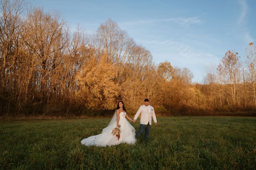 Bride and groom holding hands in an open field with autumn trees behind them