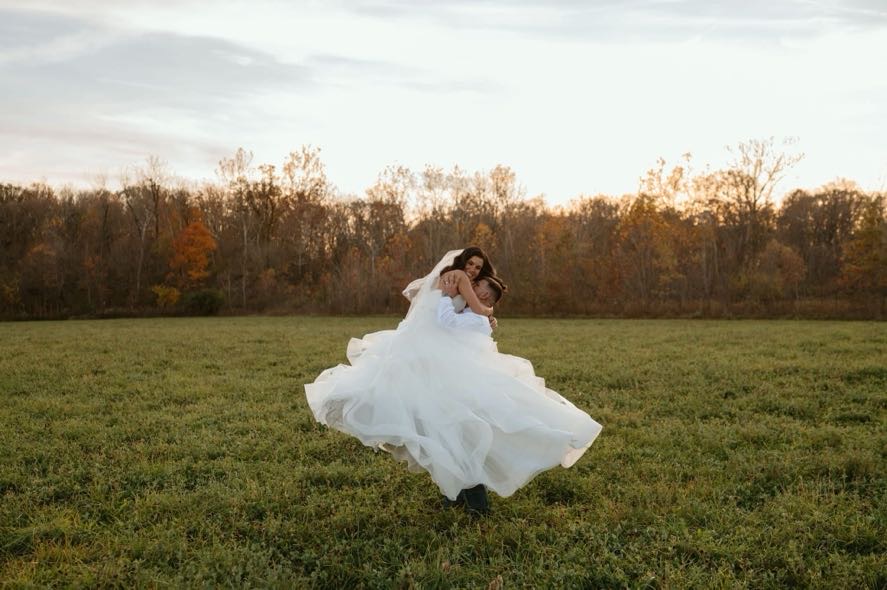 Groom lifting bride joyfully in a field surrounded by autumn colors during their fall foliage wedding