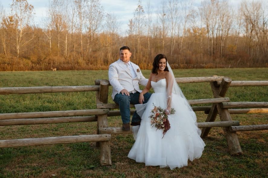Bride and groom posing with rustic charm on a split-rail fence during their fall foliage wedding