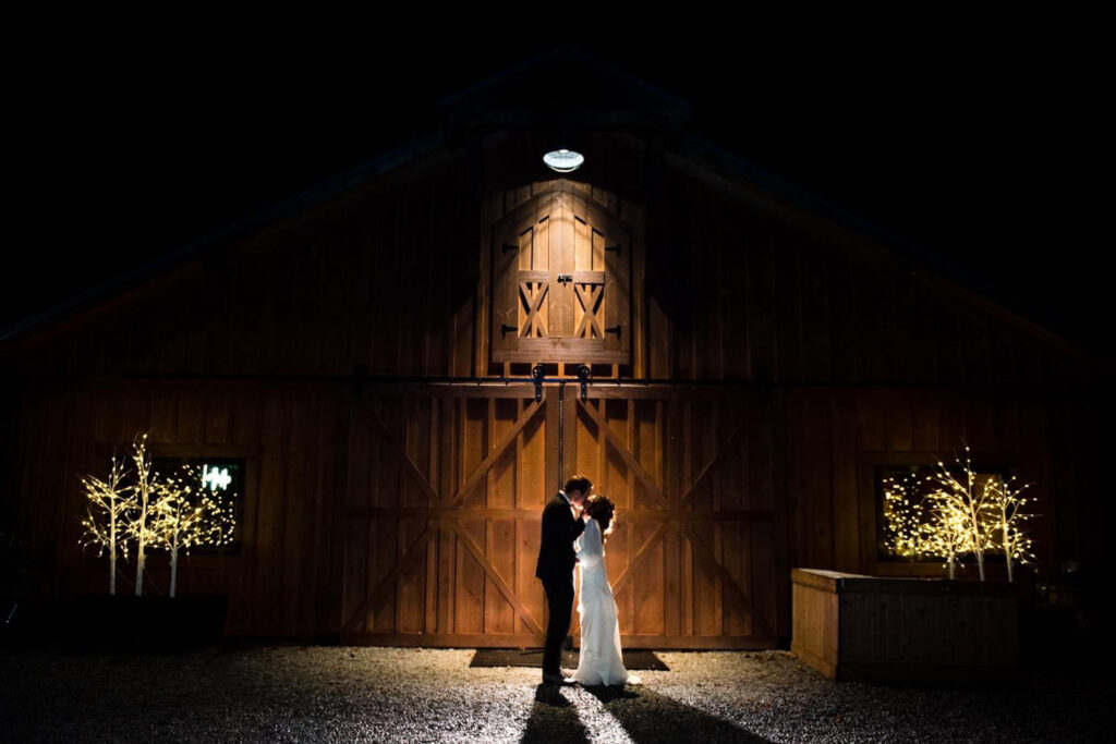 A bride and groom are kissing in front of a barn.