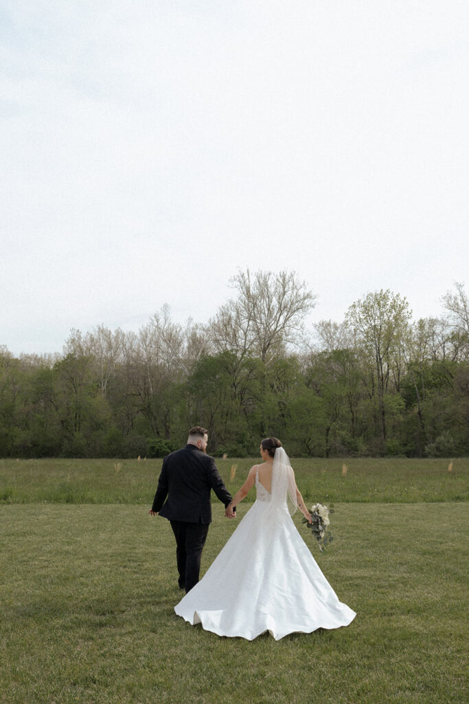 A bride and groom are walking through a grassy field holding hands.