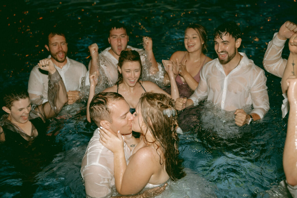 Newlywed couple kissing in a pool surrounded by cheering friends