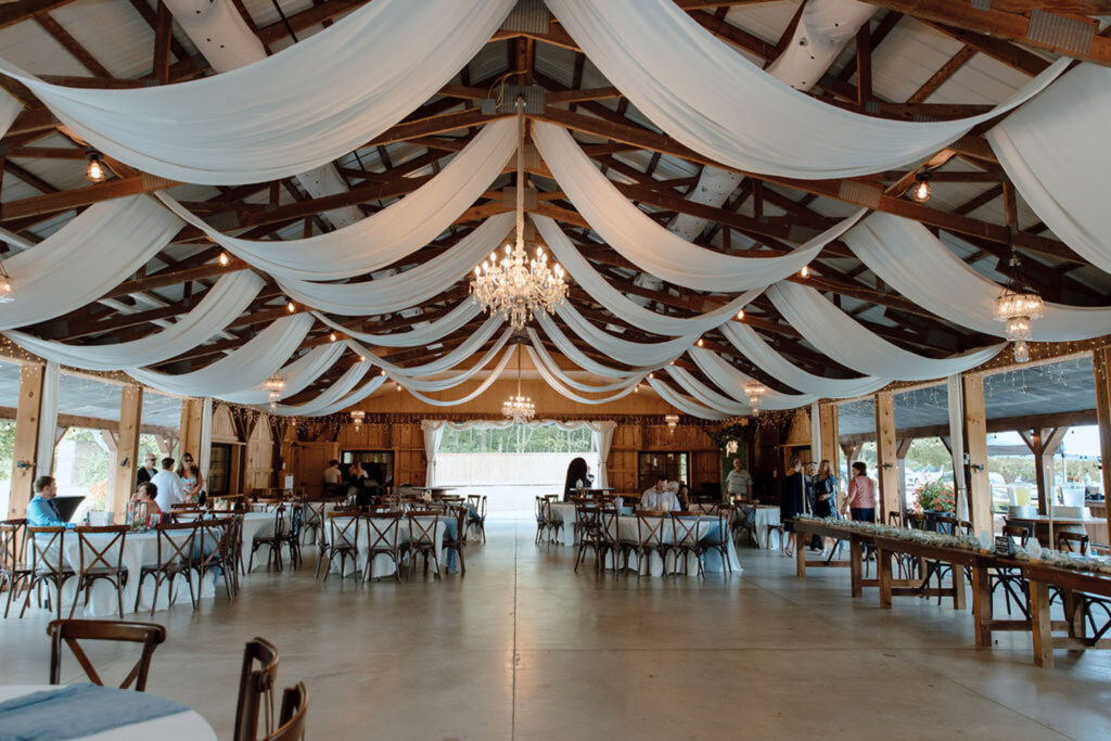 Wide interior view of a barn wedding venue featuring chandeliers and white draping, offering a glimpse of what’s included in venue pricing for weddings