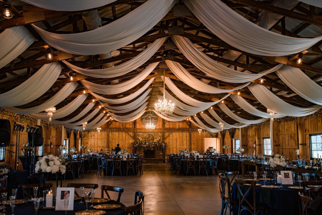 Rustic wedding reception hall with draped fabric ceiling, chandeliers, and elegantly set tables.