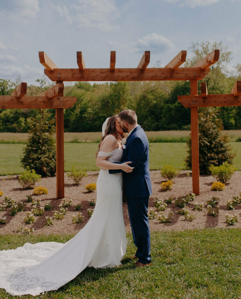 Bride and groom kissing under a wooden pergola in a grassy field.