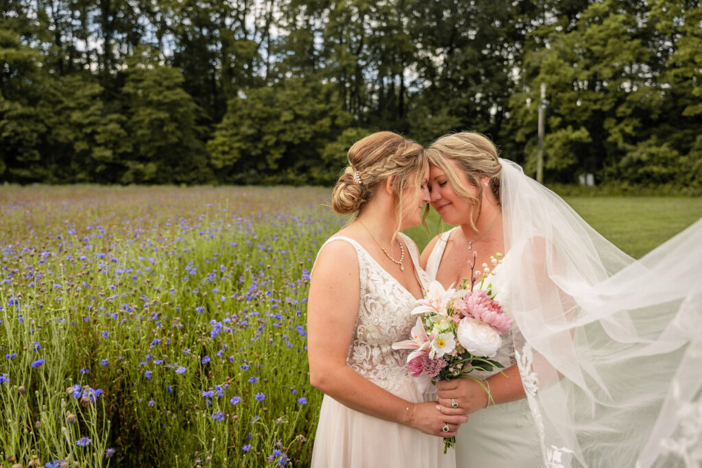 Two brides embracing in a purple wildflower meadow with bouquet and veil.