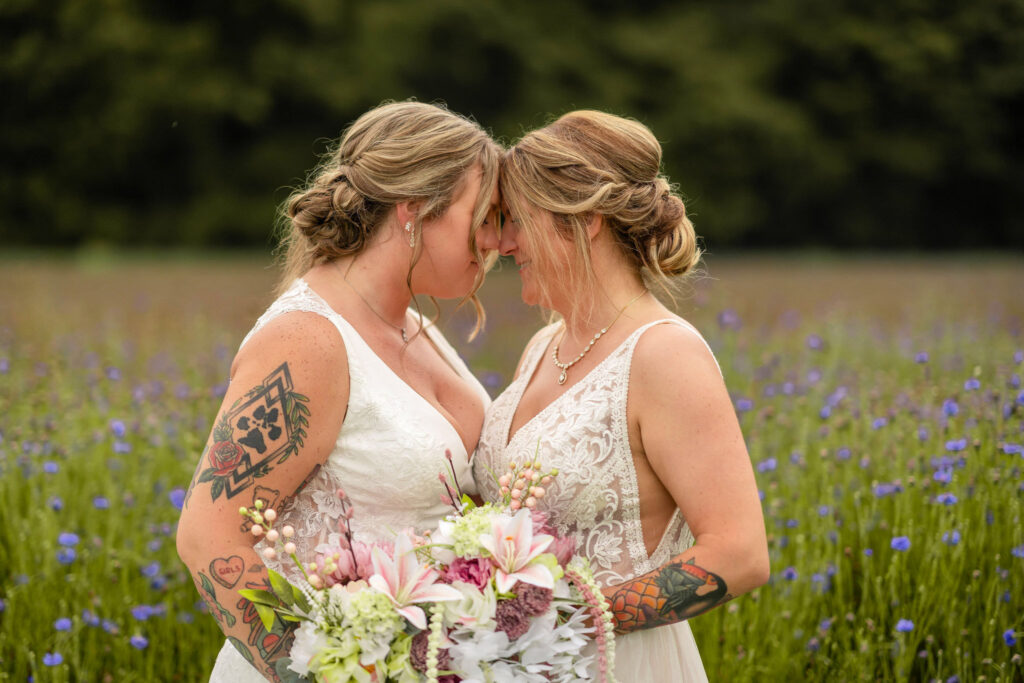 Close-up of two brides touching foreheads, holding pastel bouquets in a field.