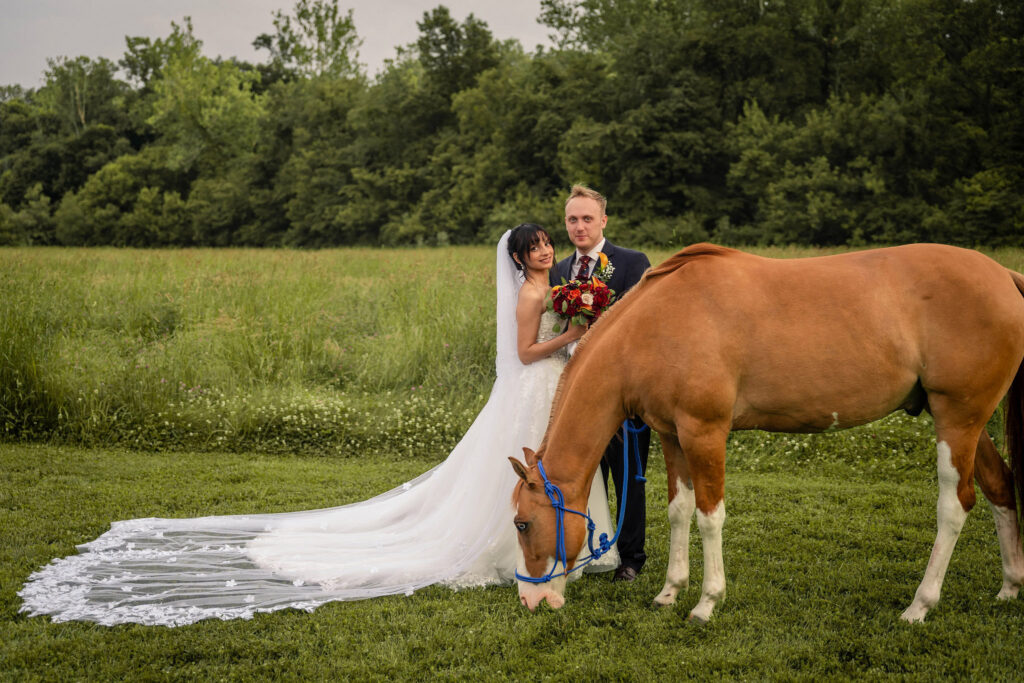 Bride and groom posing with a horse grazing in a meadow.