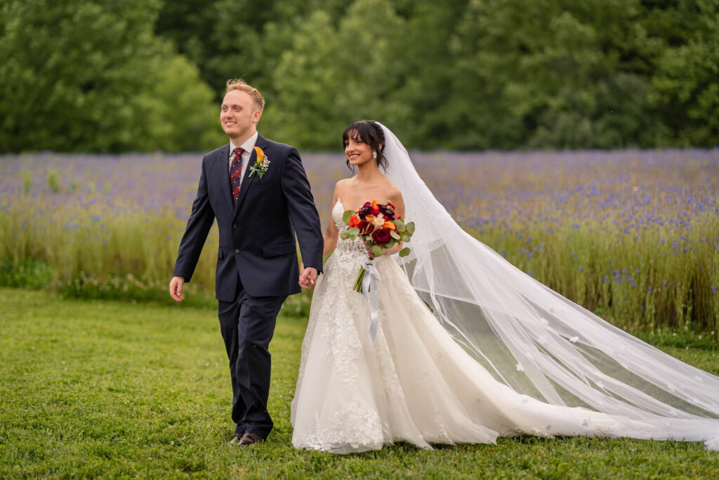 Bride and groom walking hand in hand through a wildflower field with flowing veil.