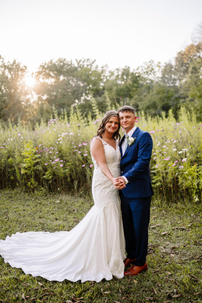 Bride and groom holding hands in front of blooming wildflowers at sunset.